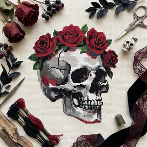 May include: A cross-stitch pattern on cream fabric featuring a human skull adorned with a crown of vibrant red roses and green leaves. The design is surrounded by dried red roses, black berries, metal scissors, a small skull bead, and thread.