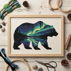 May include: A framed cross-stitch embroidery featuring the silhouette of a bear filled with a landscape of a dark forest, a reflective lake, and vibrant green aurora borealis lights against a starry night sky on a light-colored fabric background.