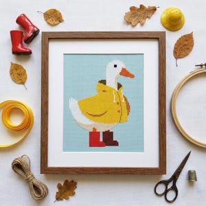 May include: A framed cross-stitch embroidery featuring a white duck wearing a yellow raincoat and mismatched red and brown boots against a light blue background. The wooden frame surrounds the white mat and the stitched artwork of the duck.