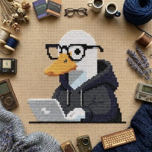 May include: A cross-stitch pattern on beige fabric featuring a white duck wearing black-rimmed glasses and a dark gray hoodie, sitting at a laptop. The design is surrounded by craft supplies including spools of thread, dried flowers, and small electronics.