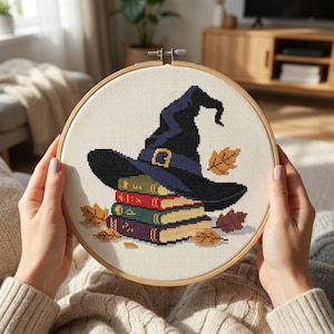 May include: A circular wooden embroidery hoop containing a cross-stitch design of a dark blue witch hat resting on a stack of four colorful books, surrounded by scattered orange and brown autumn leaves on a cream-colored fabric background.