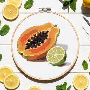 Könnte beinhalten: Kreuzstich-Stickerei einer halben Papaya und einer Limettenscheibe, in einem Holzreifen. Die Papaya ist orange mit schwarzen Samen, und die Limette ist grün. Zitronenscheiben und Minzblätter umgeben den Reifen auf einer weißen Oberfläche.