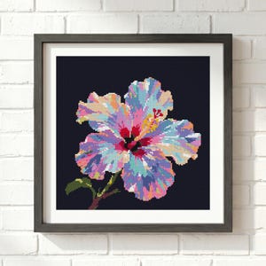 May include: A square framed wall art piece featuring a pixelated cross-stitch pattern of a hibiscus flower. The flower displays a vibrant mix of pink, blue, purple, and yellow hues against a solid dark navy background, set in a dark wood frame.