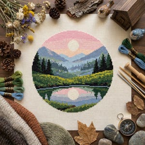May include: A circular cross-stitch embroidery featuring a landscape with a pink sky, a pale sun, blue mountains, dark green pine trees, and a grassy field reflected in a calm lake, surrounded by dried flowers, pinecones, and embroidery floss.