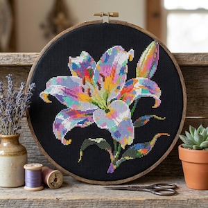 May include: A circular wooden embroidery hoop containing a cross-stitch pattern of a multi-colored lily flower on a black fabric background. The lily features vibrant shades of pink, purple, yellow, blue, and white, with a single bud to the right.