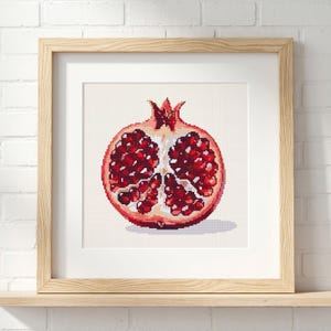 Pode incluir: Obra de arte em ponto cruz de uma romã cortada ao meio, mostrando sementes vermelhas vibrantes e uma casca de cor clara. A obra está emoldurada em uma moldura de madeira clara e é exibida em uma prateleira de madeira contra uma parede de tijolos brancos.