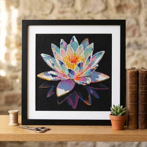 May include: A square framed cross-stitch artwork featuring a vibrant, multi-colored lotus flower with petals in shades of pink, blue, and yellow against a solid black background. The piece is displayed in a simple black frame with a white mat.