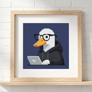 May include: A framed pixel art print featuring a white duck with an orange beak wearing black-rimmed glasses and a dark gray hooded sweatshirt. The duck is positioned in front of a light gray laptop against a solid dark blue background.