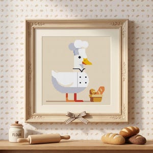 May include: A framed cross-stitch pattern featuring a white duck wearing a chef hat and a white chef jacket with black buttons. The duck stands next to a small brown basket filled with orange and brown bread loaves on a light beige background.