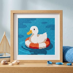 May include: A framed cross-stitch embroidery featuring a white duck with a yellow beak floating in blue water inside a red and white striped life preserver. The artwork is encased in a light-colored wooden frame with a white mat border.