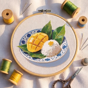 May include: A cross-stitch embroidery project in a wooden hoop featuring a plate with a sliced yellow mango and a mound of white sticky rice on green leaves. The plate has a blue and white floral pattern. Spools of thread, needles, and a thimble surround it.