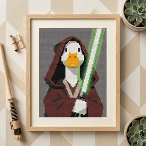 May include: A framed cross-stitch artwork featuring a pixelated white goose with an orange beak wearing a dark red hooded robe. The goose holds a glowing green light saber. The design is set against a gray background and enclosed in a light wood frame.