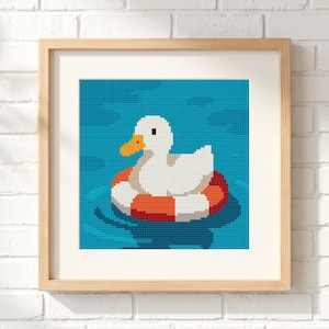 May include: A square framed cross-stitch artwork featuring a white duck with an orange beak floating in blue water inside a red and white striped life preserver. The piece is encased in a light-colored wooden frame with a clean white mat border.