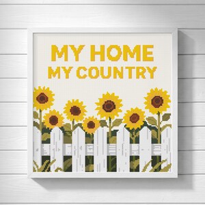 May include: A square cross-stitch pattern in a white frame featuring yellow sunflowers behind a white picket fence. The text MY HOME MY COUNTRY is stitched in yellow block letters at the top against a cream-colored background.