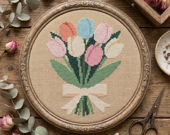 May include: A circular wooden embroidery hoop containing a cross-stitch pattern of a tulip bouquet. The design features peach, light blue, pink, and white tulips with green stems and leaves, tied with a cream-colored ribbon on tan fabric.