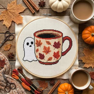 Puede incluir: Un proyecto de bordado en punto de cruz en un bastidor de madera circular que muestra una taza color crema llena de líquido oscuro, decorada con hojas de otoño naranjas, rojas y marrones, junto a un pequeño fantasma blanco con dos ojos negros.
