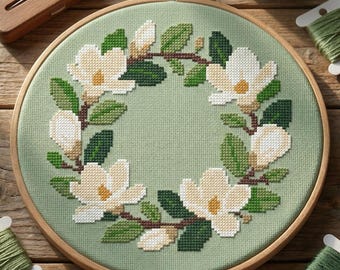 May include: A circular wooden embroidery hoop containing a cross-stitch pattern of a wreath featuring white magnolia flowers with yellow centers and dark green leaves on a light sage green fabric background, surrounded by embroidery floss.