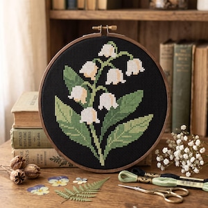 May include: A circular wooden embroidery hoop containing a cross-stitch pattern of white lily of the valley flowers with green leaves on a black fabric background. The design features multiple bell-shaped blossoms and broad, pointed foliage.