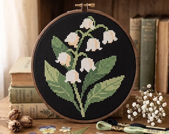 May include: A circular wooden embroidery hoop containing a cross-stitch pattern of white lily of the valley flowers with green leaves on a black fabric background. The design features multiple bell-shaped blossoms and broad, pointed foliage.