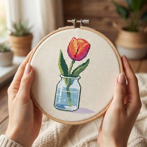 May include: A circular wooden embroidery hoop containing a cross-stitch pattern on cream-colored fabric. The design features a single red and orange tulip with green leaves, placed inside a clear glass jar filled with water, showing a light blue tint.
