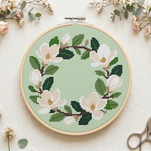 May include: A cross-stitch embroidery hoop featuring a magnolia flower wreath design. The flowers are white with yellow centers, surrounded by green leaves and branches, set against a light green background. The wooden hoop is visible, and the piece is displayed on a white surface.