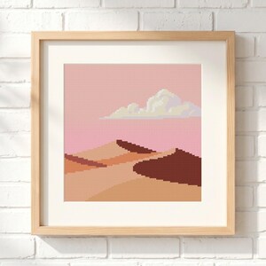 Peut inclure: Une broderie au point de croix carrée représentant un paysage désertique avec des dunes de sable dans des tons de beige, orange et brun foncé sous un ciel rose uni avec un nuage blanc et jaune clair, encadrée dans un cadre en bois clair.