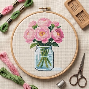 May include: A cross-stitch project in a wooden embroidery hoop featuring a bouquet of pink peonies in a glass mason jar. The kit includes pink and green embroidery floss, a wooden needle case with needles, and metal embroidery scissors.