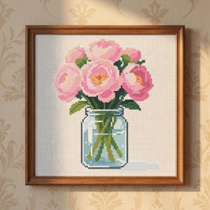 May include: A square cross-stitch embroidery featuring a bouquet of pink peonies in a clear glass mason jar. The flowers have shades of light and dark pink with yellow centers, and the stems are green. The artwork is set in a wooden frame.