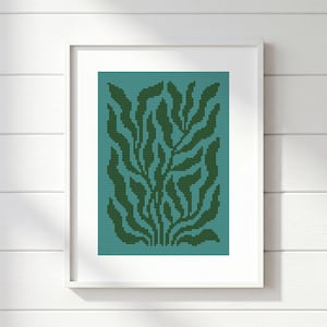 May include: A framed cross-stitch artwork featuring a stylized botanical design. The artwork showcases a pattern of dark green leaves against a teal background. The frame is white, and the piece is displayed against a white wooden wall.