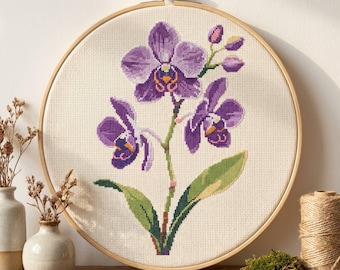 May include: A circular wooden embroidery hoop containing a cross-stitch pattern of a purple orchid plant with two large blooms, one smaller bloom, and two closed buds, set against a cream-colored fabric background with green leaves.