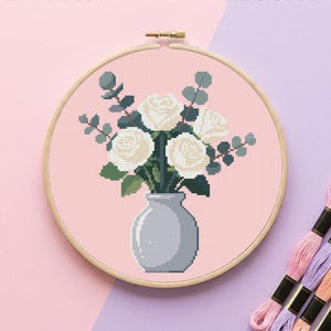 Könnte beinhalten: Kreuzstich-Stickerei mit weißen Rosen und Eukalyptus in einer grauen Vase, vor rosa Hintergrund. Das Design ist in einem Holzreifen gerahmt. Spulen mit Stickgarn in Rosa- und Lilatönen sind sichtbar.