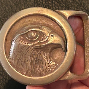 May include: A circular brass belt buckle featuring a raised relief design of an eagle head in profile. The metallic surface has a warm, burnished tone with detailed engravings of the bird's feathers and eye, set within a smooth, polished frame.