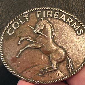 May include: An oval-shaped metal belt buckle with a rope-style border. The center features a raised relief of a rearing horse holding a spear, positioned above a globe grid. The text COLT FIREARMS is embossed in capital letters along the top edge.
