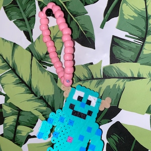 Puede incluir: Un llavero de cuentas azul claro y rosa con un diseño de personaje de dibujos animados pixelado. El personaje tiene una cara sonriente, dos ojos y dos brazos. El llavero está unido a una pulsera de cuentas rosas.