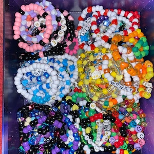 May include: A box filled with colorful beaded bracelets. The bracelets are made with small plastic beads and have different words and designs. Some of the bracelets have smiley faces, hearts, and stars.