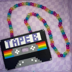 May include: A black cassette tape necklace with the word "TAPE" in white and purple. The cassette tape has a rainbow design and is connected to a beaded necklace with rainbow-colored beads.