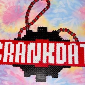 May include: A handmade, pixelated sign with the word "CRANKDAT" in white block letters on a red background. The sign is attached to a black and red beaded chain, with a black and red geometric design. The background is a tie-dye pattern.