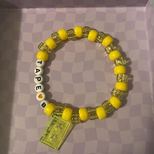 Pode incluir: Uma pulseira de contas amarelas com a palavra "TAPE" escrita em contas de letras brancas. Uma pequena conta de coração laranja está entre o "E" e o "B". Um pingente retangular transparente pende da pulseira.