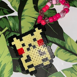 Pode incluir: Um amuleto pixelado amarelo, preto e vermelho com um bracelete de contas rosa e branco anexado. O amuleto tem a forma de um quadrado com cantos arredondados.