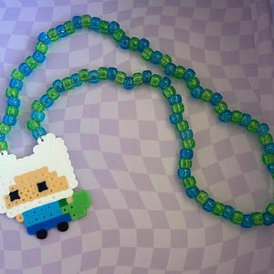 Könnte beinhalten: Eine Halskette aus blauen und grünen Perlen mit einem Anhänger, der eine pixelige Cartoonfigur darstellt. Der Anhänger hat einen weißen Hintergrund, mit einer Figur, die ein blaues Hemd und ein grünes Accessoire trägt.