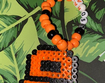 May include: Orange and black beaded bracelet with the numbers 5074 and the word "TAM" spelled out on the beads. A black and orange pixelated hat charm is attached to the bracelet.