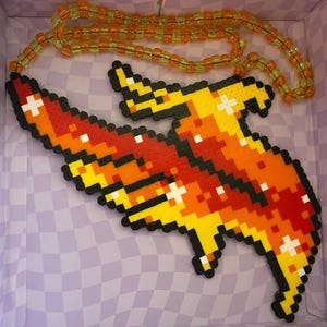 May include: A pixelated pendant necklace featuring a flame design in shades of red, orange, and yellow, with white cross accents. The necklace has a beaded chain in orange and yellow. The pendant is made of small, colorful beads.