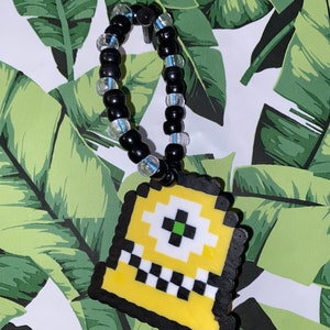 Pode incluir: Um amuleto de contas amarelas e pretas com um design pixelado de um táxi. O amuleto está preso a uma pulseira de contas pretas e brancas.