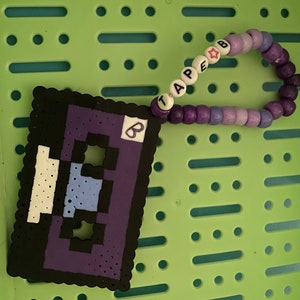 May include: A purple and black pixel art cassette tape charm with a white star and the word "tape" spelled out in white beads on a purple beaded bracelet.