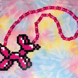 Könnte beinhalten: Eine rosa Perlenkette mit einem pixeligen Hundeanhänger. Der Hundeanhänger besteht aus rosa, schwarzen und weißen Perlen. Die Kette befindet sich auf einem Hintergrund mit Batikmuster in Rosa, Blau und Gelb.