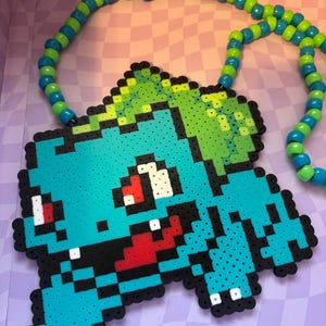May include: A handmade necklace featuring a pixelated Bulbasaur pendant made of colorful beads. The pendant is primarily teal, green, black, and red. The necklace chain is made of alternating blue and green beads. The necklace is displayed on a purple and white checkered surface.
