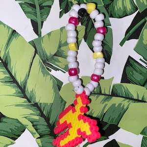 Puede incluir: Un brazalete de cuentas blancas, rosas y amarillas con un dije de avión pixelado amarillo y rojo.