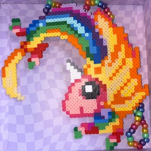 Könnte beinhalten: Ein farbenfrohes Einhorn aus geschmolzenen Kunststoffperlen. Das Einhorn hat eine Regenbogenmähne und -schwanz, einen rosa Körper und ein weißes Horn. Das Design befindet sich vor einem karierten Hintergrund, mit einer aufgefädelten, regenbogenfarbenen Schlaufe.