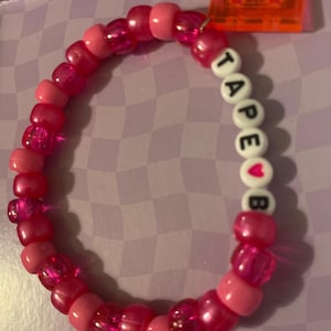 Pode incluir: Uma pulseira de contas rosa com a palavra "TAPE" e um símbolo de coração seguido da letra "B" escrita com contas de letras brancas. A pulseira é feita de várias tonalidades de contas rosa e tem um berloque de fita cassete laranja.