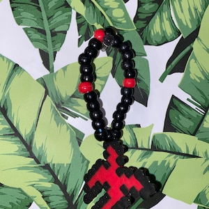 Puede incluir: Un brazalete de cuentas negras y rojas con un dije de cruz roja. El brazalete está hecho de pequeñas cuentas negras con acentos rojos. El dije de cruz está hecho de cuentas rojas y negras y tiene la forma de una cruz.